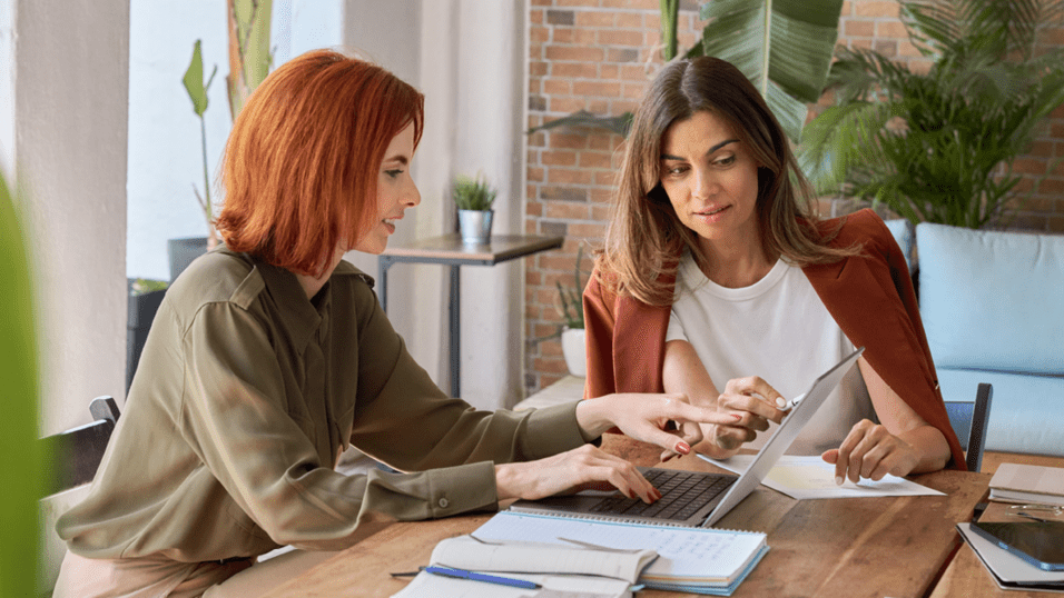 Zwei Frauen sitzen an einem Holztisch, arbeiten am Laptop und diskutieren, umgeben von Pflanzen und moderner Einrichtung.